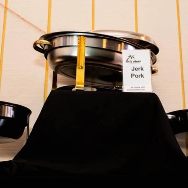 A chafing dish with "Jerk Pork" label from Irie Diner Caribbean Restaurant on a black tablecloth.