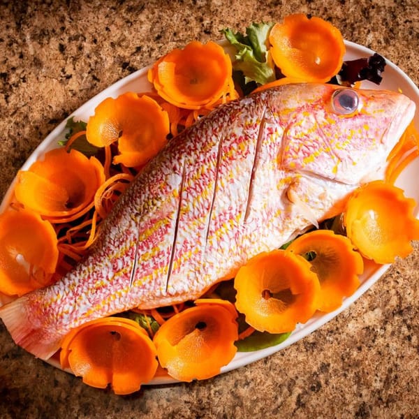 Whole roasted fish, scored and garnished with orange peels and shredded carrots on a white platter.