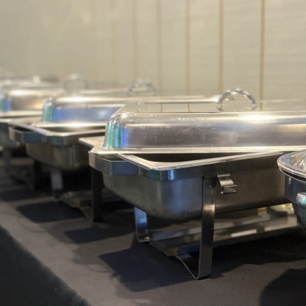 A row of silver chafing dishes with lids on a black tablecloth, ready for a buffet.