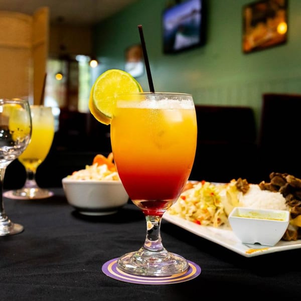 A vibrant red and orange cocktail with a lime slice and straw, next to Jamaican food.