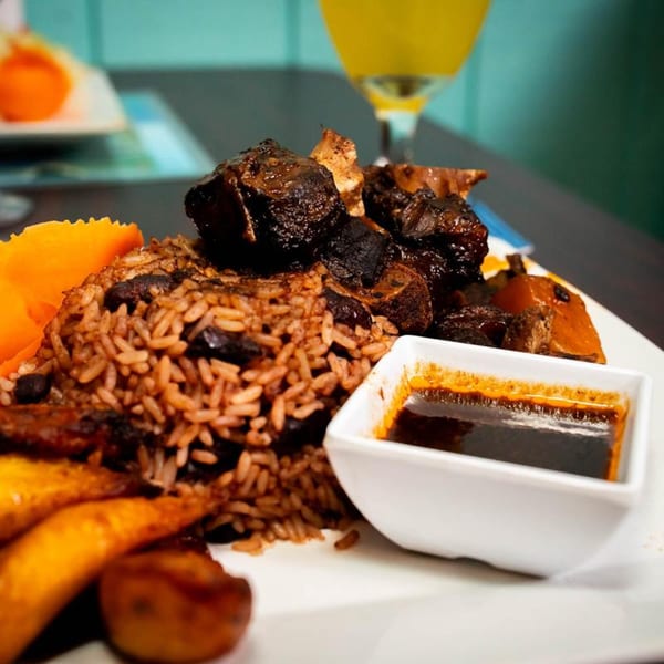 A Jamaican dish with brown rice, dark meat, plantains, and a side of sauce.