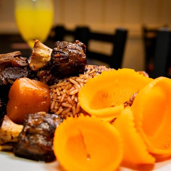 Close-up of Jamaican oxtail stew with rice and peas, plantains, and a drink in the background.