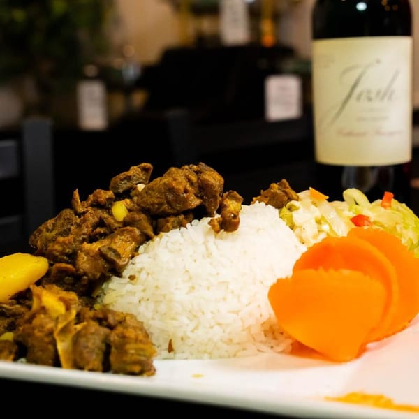 A plate of Jamaican curry goat with white rice, plantains, and coleslaw, with a wine bottle in the background.