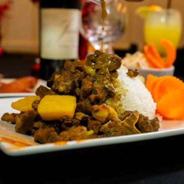 Curried goat with rice and potatoes, garnished with carrots, and a drink in the background.