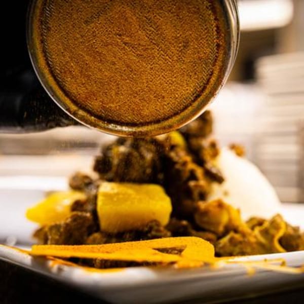 Curry powder being sprinkled from a shaker onto a plate of Jamaican curry chicken with rice.