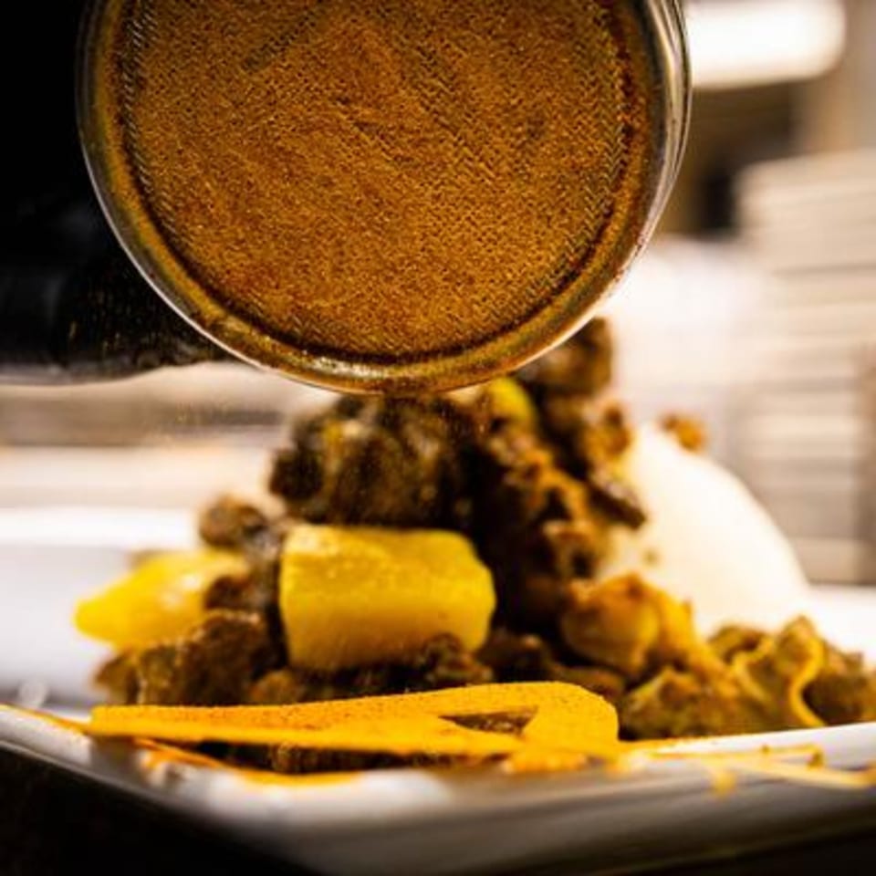 Curry powder being sprinkled from a shaker onto a plate of Jamaican curry chicken with rice.
